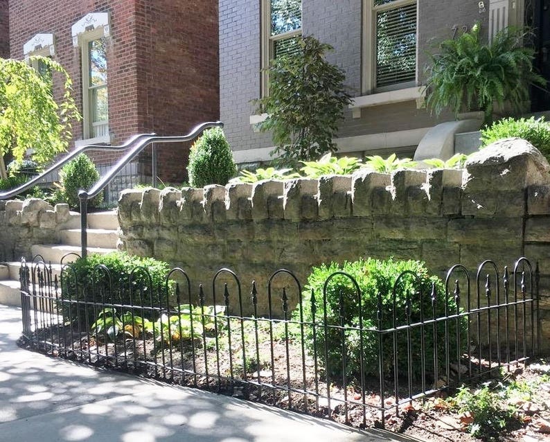 May include: A black metal fence with rounded top rails surrounds a small garden area in front of a brick building. The fence is in front of a stone wall and green bushes.
