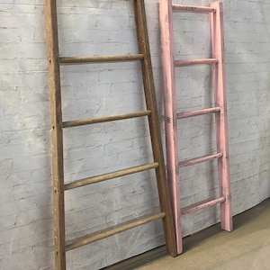 May include: Two wooden ladders, one natural wood and one painted pink, leaning against a white brick wall.