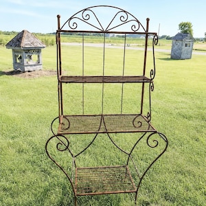 May include: A three-tier metal shelving unit with a rusted finish. The structure features a decorative arched top with scrollwork, wire mesh shelves, and curved legs. The frame is constructed from thin metal rods with a weathered, brown texture.