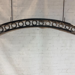 May include: A black metal arched decorative piece with circular accents, suspended against a white brick wall. The design features a series of evenly spaced rings within the curved frame, creating a unique visual element. The piece is likely for interior or exterior decor.