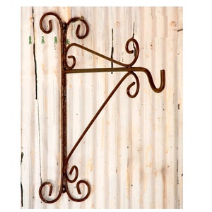 May include: Rustic metal plant hanger with a decorative scroll design. The hanger is attached to a white wooden wall.
