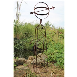 May include: A rusty metal garden sculpture with a sphere and arrow on top of a tall, square base. The sculpture is in a garden setting with green plants and rocks.