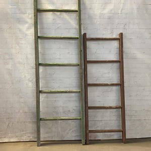 May include: Two wooden ladders leaning against a white brick wall. The ladder on the left is green and the ladder on the right is brown.
