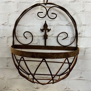 May include: A wall-mounted metal basket with a rusted brown finish. The structure features a semi-circular base with wire detailing, a flat horizontal band, and an upper arched frame decorated with symmetrical metal scrolls and a central fleur-de-lis.