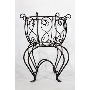 May include: A wrought iron plant stand with a round basket and decorative scrollwork. The stand has a dark brown finish and is supported by curved legs. The basket has handles and a decorative pattern. This vintage piece is suitable for indoor or outdoor use.