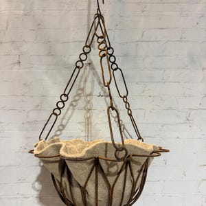 May include: A hanging basket featuring a dark brown, rusted metal wire frame with a scalloped rim. The basket is suspended by three matching rusted metal chains that meet at a central hook. A beige, felt-like fabric liner sits inside the wire frame.