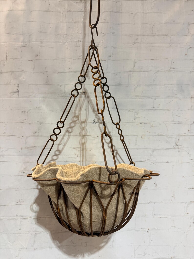 May include: A hanging basket featuring a dark brown, rusted metal wire frame with a scalloped rim. The basket is suspended by three matching rusted metal chains that meet at a central hook. A beige, felt-like fabric liner sits inside the wire frame.