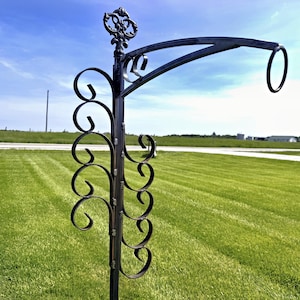 May include: A black wrought iron hanging plant stand with decorative scrollwork and a circular ring. The stand is set against a backdrop of a green lawn and a blue sky.
