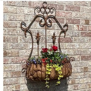 May include: A rusty metal wall planter with a decorative scrolled design. The planter is filled with red, pink, and white flowers and green foliage.