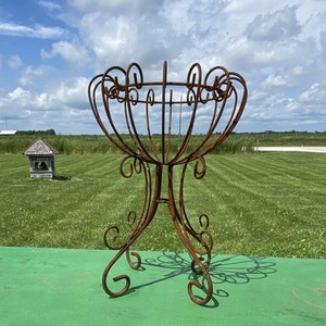 May include: A tall, ornate, rusty brown metal plant stand with a decorative, openwork basket design. The stand has a three-legged base with scrolled details and is set against a green background under a cloudy sky.