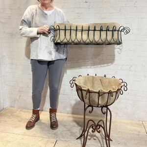 May include: A rectangular black metal planter with a tan fabric liner and a matching round metal planter on a tall, ornate stand with a tan fabric liner. The rectangular planter features decorative scrollwork along the top edge.