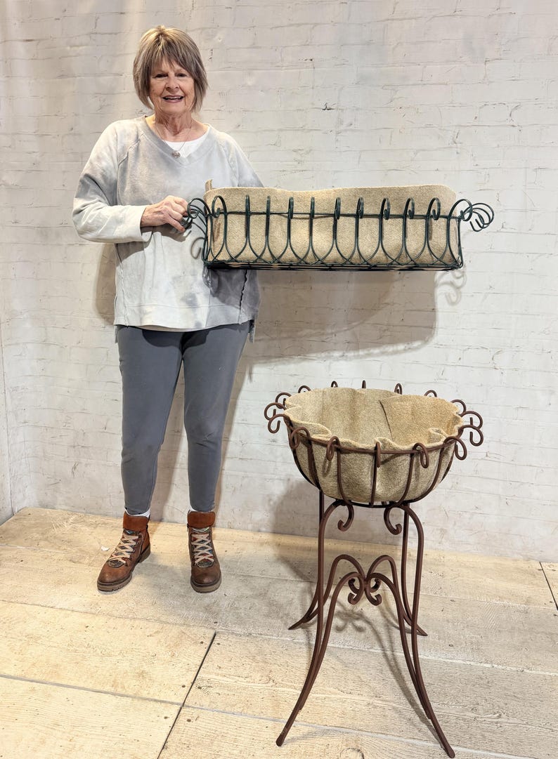 May include: A rectangular black metal planter with a tan fabric liner and a matching round metal planter on a tall, ornate stand with a tan fabric liner. The rectangular planter features decorative scrollwork along the top edge.