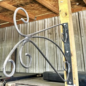 May include: A handcrafted metal plant hanger with decorative scrollwork. The gray metal hanger is mounted on a wooden post with a black metal bracket. The design features curved lines and swirls, creating an elegant and artistic look.