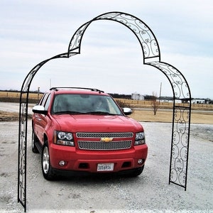 May include: A red SUV is parked under a black wrought iron garden arch. The arch has decorative scrollwork and is set on a gravel driveway. The car has a chrome grill and side mirrors.