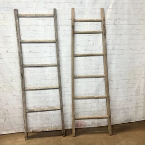 May include: Two wooden ladders leaning against a white brick wall. The ladders are made of natural wood and have five rungs each.