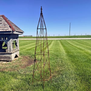 May include: A tall, rusty metal garden obelisk stands on a grassy lawn next to a weathered wooden gazebo. The obelisk has a pointed top and a lattice design. The sky is clear blue.