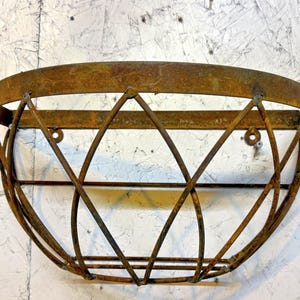 May include: A rustic, wall-mounted metal planter with a weathered, brown finish. The planter features a semi-circular top and a geometric, diamond-patterned cage design. Includes two mounting holes for easy installation.