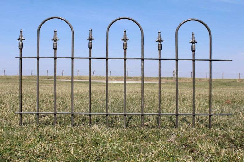 May include: A black metal fence with arched tops and spear-shaped finials. The fence is in a grassy field with a blue sky in the background.