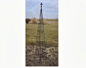 Op de afbeelding: Een hoge tuinobelisk van donkerbruin metaal met een vierzijdig piramideframe opgebouwd uit dunne verticale en horizontale staven. De structuur loopt taps toe naar een punt aan de bovenkant, afgewerkt met een decoratieve fleur-de-lis knop.