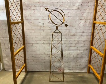 Puede incluir: Un obelisque de jardín de metal dorado con un diseño decorativo de flecha y círculo en la parte superior. El obelisco está hecho de finas barras de metal que crean un patrón de diamante.