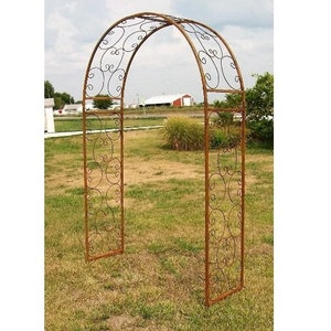 May include: A rusty brown metal garden arch with a decorative scrolled design. The arch is standing in a grassy field.