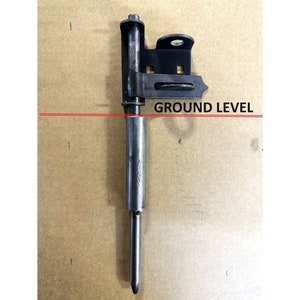 May include: A black metal gate latch with a long, pointed metal rod. The rod is attached to a metal plate with a hole for a screw. The latch is designed to be mounted to a gate post and the rod is inserted into the ground.  GROUND LEVEL is written on the image.