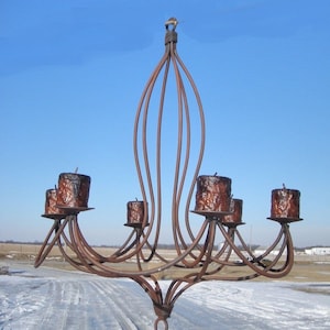 May include: A rustic metal chandelier with five candle holders. The chandelier is made of dark brown metal and has a simple, elegant design. The candle holders are made of a similar metal and are designed to hold pillar candles.