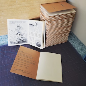 May include: A stack of 73 brown paperbacks with the title "Sharbala" on the top book. The book is open to a page with a black and white illustration of a person sitting on a stool. The text on the page reads "We'd eat ice cold sandwiches by the side of the road. White bread, ham, mayonnaise and we saved money."