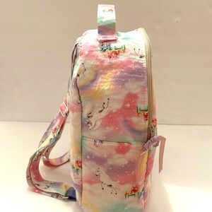 May include: A white backpack with a rainbow and unicorn print. The backpack has a zipper closure and a top handle.