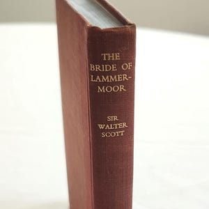May include: A hardcover book titled "The Bride of Lammermoor" by Sir Walter Scott. The book has a burgundy cover with gold lettering. The book is standing upright on a white surface.