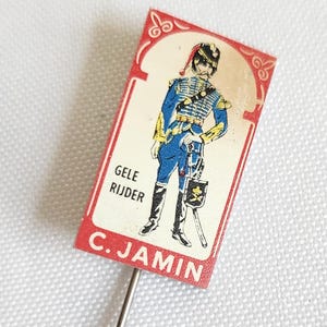 May include: A vintage pin featuring a soldier in a blue uniform with gold accents, standing on a red rectangular background. The pin reads "GELE RUIJER" and "C. JAMIN". The pin is set against a white background.