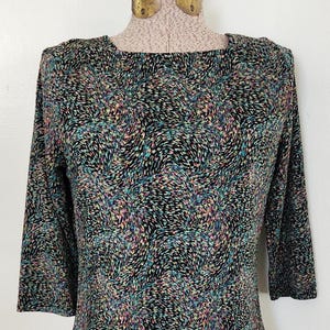 May include: A three-quarter sleeve top with a colorful, abstract pattern. The top features a black background with a design of small, multicolored shapes in shades of teal, pink, and peach. The top is displayed on a mannequin.