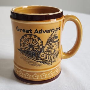 May include: A tan ceramic mug with a brown rim and handle. The mug features a black illustration of a Ferris wheel, train, and the words "Great Adventure". The mug has a decorative base.