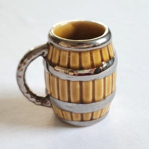 May include: A miniature mug shaped like a barrel, with a yellow body and silver-colored metal bands and handle. The mug has a small, round shape and is likely a decorative item.