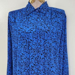 May include: A long-sleeved button-down shirt in a vibrant blue hue, adorned with a black floral pattern. The shirt features a classic collar and is displayed on a mannequin. The fabric appears to be lightweight.