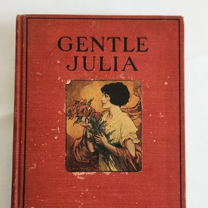 May include: A red hardcover book titled "Gentle Julia" by Booth Tarkington. The cover features an illustration of a woman in a white dress holding a bouquet of flowers.