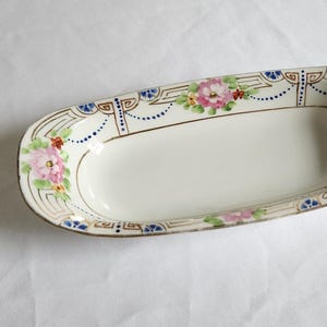 May include: An oval, off-white ceramic serving dish with a floral design. The dish is trimmed with gold and features pink flowers, blue accents, and a decorative border. A vintage piece for serving food.