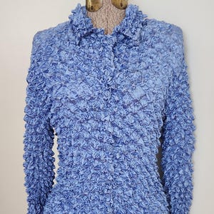 May include: A light blue textured button-down shirt with long sleeves. The shirt is displayed on a mannequin. The fabric has a unique, bubbly texture. The collar is a classic style.