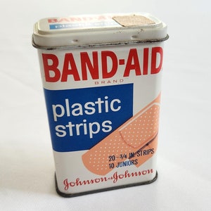 May include: A vintage tin of Band-Aid Brand plastic strips. The tin is white with blue and red lettering and a picture of a bandage. The text on the tin reads "20 - 3/4 in. Strips 10 Juniors Johnson & Johnson".