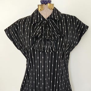 May include: Black and white striped blouse with a bow tie collar. The short-sleeved top features vertical white lines against a black background. The blouse is displayed on a mannequin.