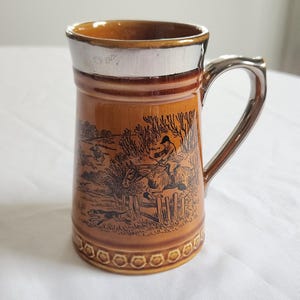 May include: A brown ceramic mug with a silver rim and handle. The mug features a black and white illustration of a hunting scene with a group of people and dogs.