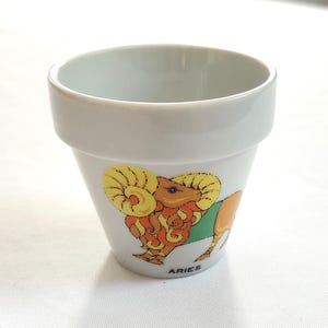 May include: A small, white ceramic pot with a flared rim. The pot features a colorful illustration of a ram, the Aries zodiac symbol, with the word "ARIES" printed below. The pot is likely used for small plants or as a decorative item.