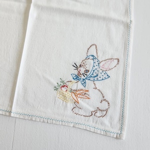 May include: White cotton tea towel with blue embroidered edging. A white rabbit with a blue and white bonnet is embroidered in the corner, holding a basket of fruit.