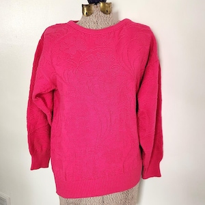 May include: A bright pink sweater with a crew neck and long sleeves. The sweater is displayed on a mannequin. The knitwear has a textured pattern across the front and sleeves. The garment is made of a soft, warm material.