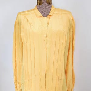 May include: A long-sleeved, yellow blouse with vertical pleats down the front. The blouse has a collared neckline and button closures. The garment is displayed on a mannequin against a white wall.
