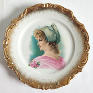 May include: A decorative porcelain plate with a gold-colored, ornate rim. The center features a portrait of a woman in profile, wearing a green hat and pink shawl. The plate has a white background and is likely vintage.