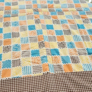 May include: A round tablecloth with a patchwork design in shades of yellow, orange, blue, and brown. The tablecloth has a brown border with a white polka dot pattern.