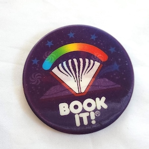 May include: A round, purple button with the text "BOOK IT!" in white. The button features a rainbow-colored design with a parachute and stars.