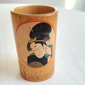 May include: A cylindrical wooden cup with a Japanese geisha illustration. The geisha is depicted in a traditional hairstyle and attire, within a circular frame. The cup has a natural wood grain finish.