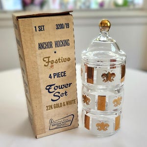 May include: A vintage Anchor Hocking Festive 4-piece tower set in its original box. The clear glass tower has gold and white fleur-de-lis designs. The box is tan with black and gold text.
